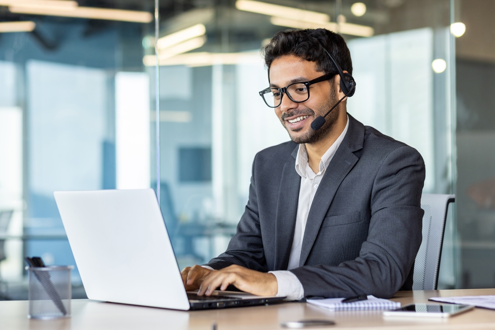 Young successful man at workplace with headset phone typing on laptop keyboard, customer support worker for online buyers inside office, hispanic helping and consulting customers.
