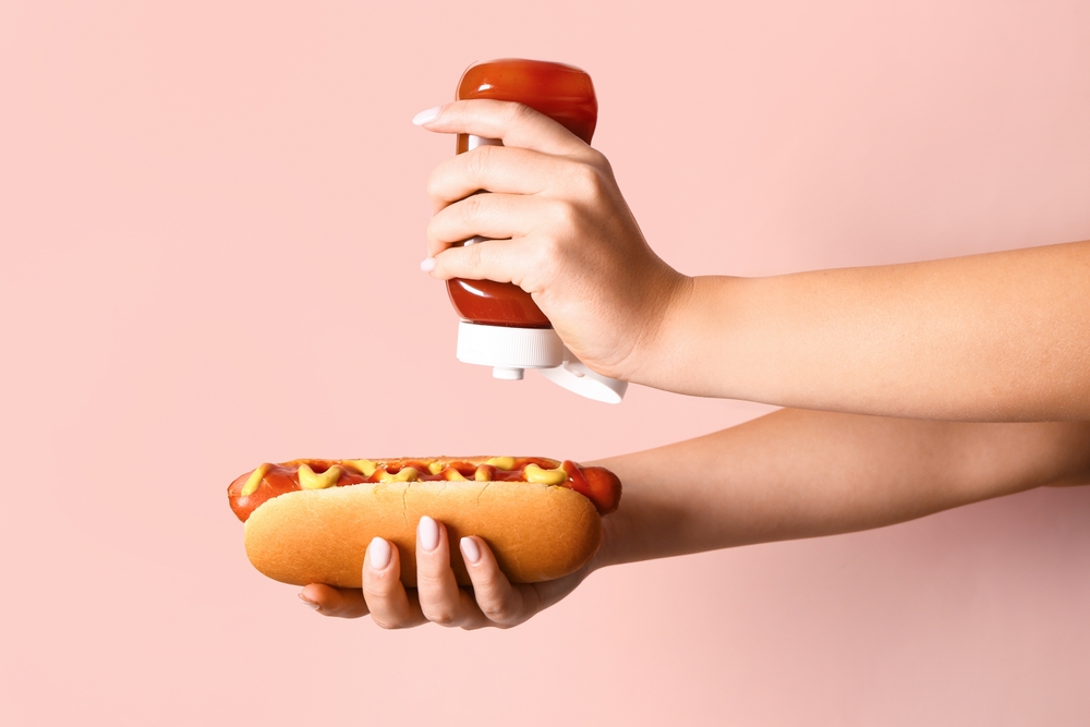 Woman adding ketchup onto tasty hot dog on pink background
