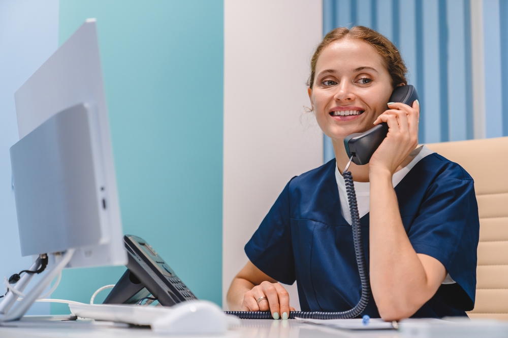 Smiling young woman nurse receptionist talking on phone while working in modern clinic. Making doctor`s appointment, consulting assisting helping customers patients clients in hospital

