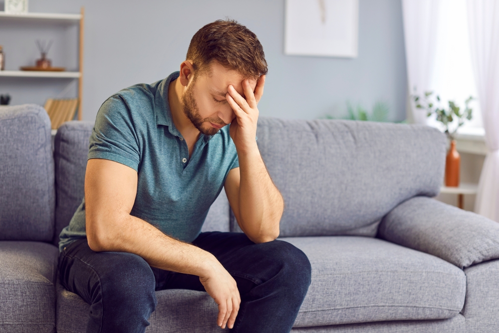 Portrait of upset sad depressed man sitting on sofa in living room at home alone suffering from headache. Young guy in bad thoughts feeling lonely indoors or having problems in relationships