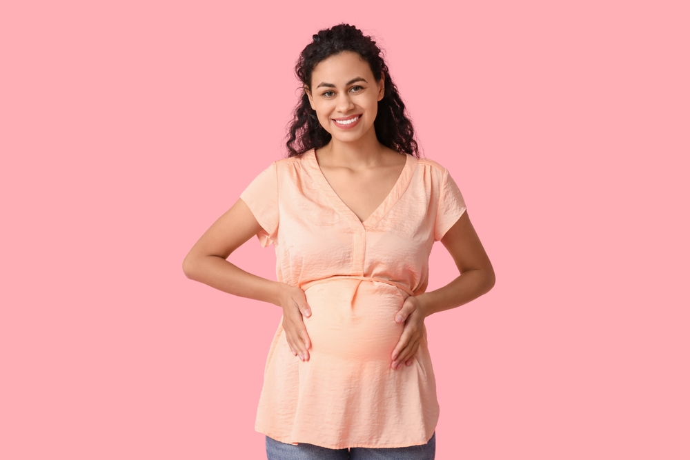 Happy young pregnant African-American woman on pink background
