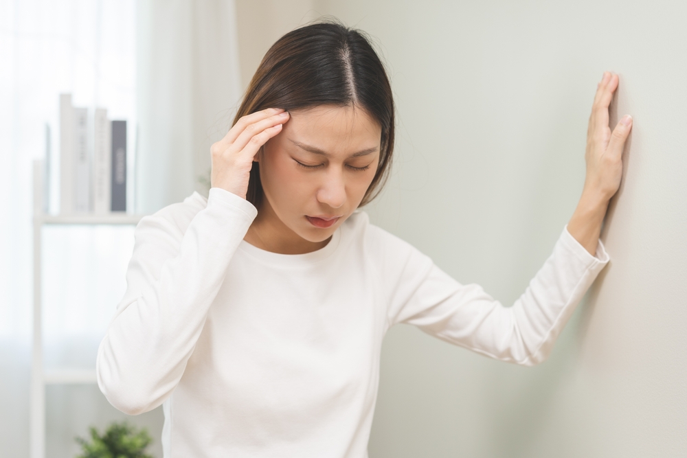 Dizzy asian young woman, girl headache or migraine pain. suffering from vertigo, difficulty standing up leaning on wall at home, holding head with hand, health problem of brain, inner ear not balance.