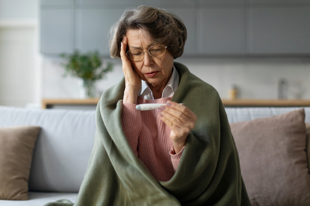 Sick elderly European woman checking her temperature, sitting wrapped in blanket on couch at home, senior lady feeling unhealthy