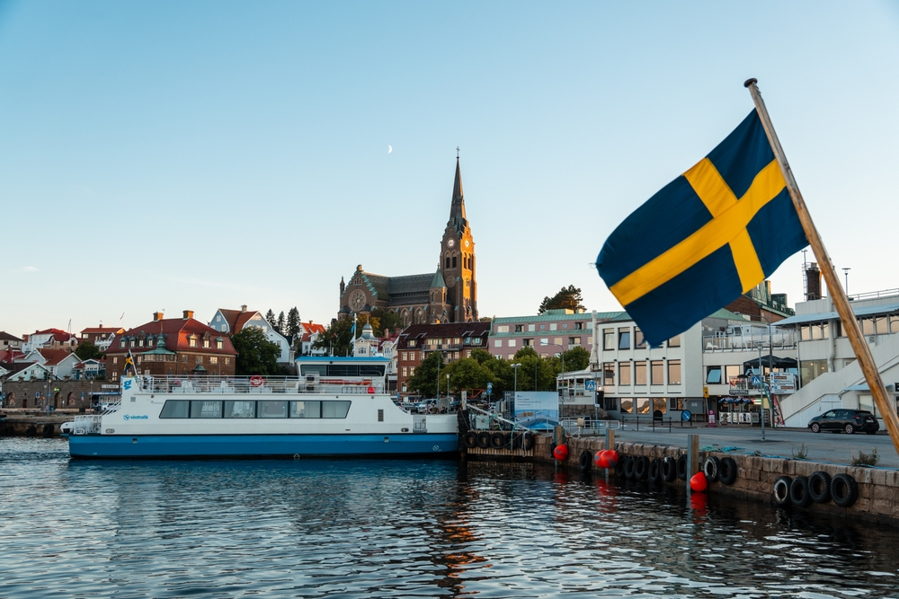 Lysekil, Sweden - July-12-2024, Ferry landing in the port with the church in the backgroundunder the moon in a midsummer night. A Sweden Flag waving in the foreground.
