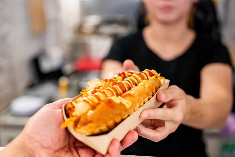 A close-up of a hand holding a hot dog with mustard and ketchup, with an out-of-focus person in the background. The hot dog is the main focus, showcasing its toppings and the casual dining experience.
