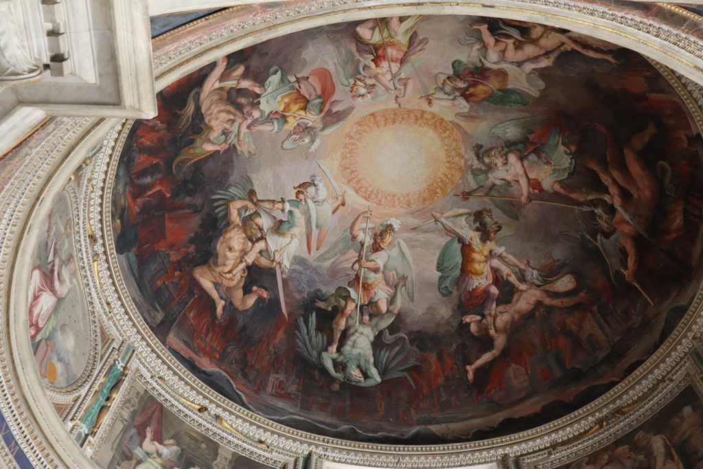 A Renaissance-style ceiling fresco in a domed church depicting angels and heavenly figures gathered around a central bright light. The painting shows winged figures with colorful garments against a backdrop of reddish flames or clouds along the edges.