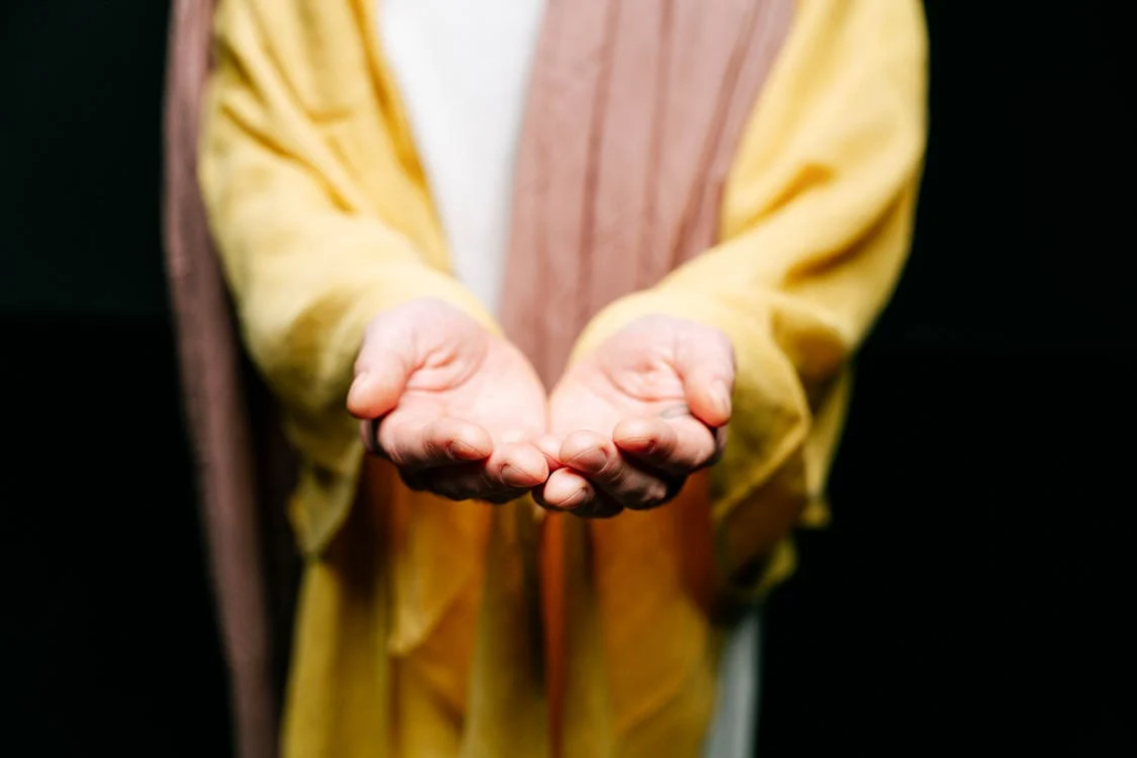 A close-up photograph of outstretched hands, palm up, wearing a yellow and burgundy robe or garment. The image is taken against a dark background and appears to represent Christ or a biblical figure offering mercy or blessing.