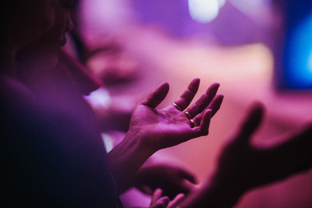 A close-up photograph of hands raised in worship, bathed in purple and blue stage lighting. The image shows just the silhouette of hands raised in prayer or praise during what appears to be a church service.