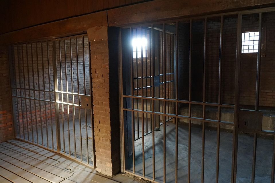 A photograph of empty prison cells viewed through iron bars. The brick-walled cells are dimly lit with light coming from small windows, showing a stark, abandoned prison interior.