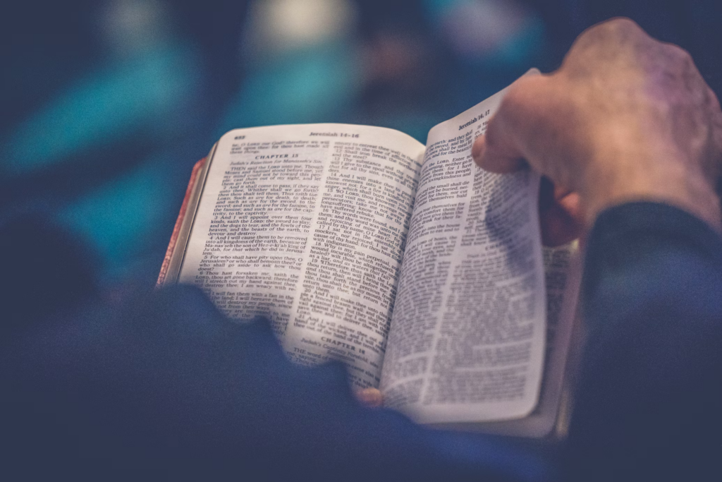  A photograph of hands holding an open Bible, with the pages visible showing small text in columns. The image is taken with a shallow depth of field with a blue background, focusing on the sacred text.