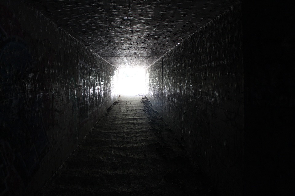 A photograph looking down a dark tunnel or passage with light visible at the end. The tunnel has stone walls and steps leading upward toward the bright opening, creating a stark contrast between darkness and light.