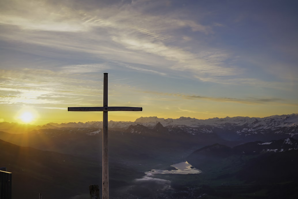  A landscape photograph of a wooden cross silhouetted against a sunset sky. The cross stands on a mountain overlook with dramatic views of mountains, a lake, and valleys below, with golden light from the setting sun illuminating the scene.