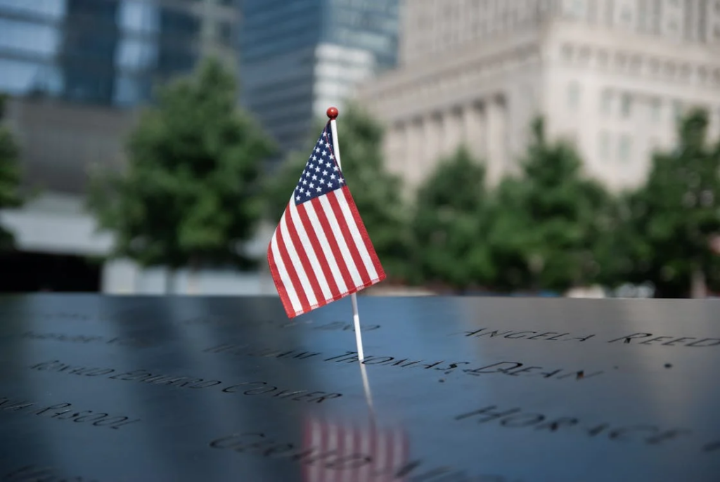 A small American flag placed in the 9/11 Memorial in New York City. The flag stands upright on a reflective black stone surface engraved with names of victims. In the background, trees and city buildings are visible but blurred, creating a solemn focus on the memorial and flag.