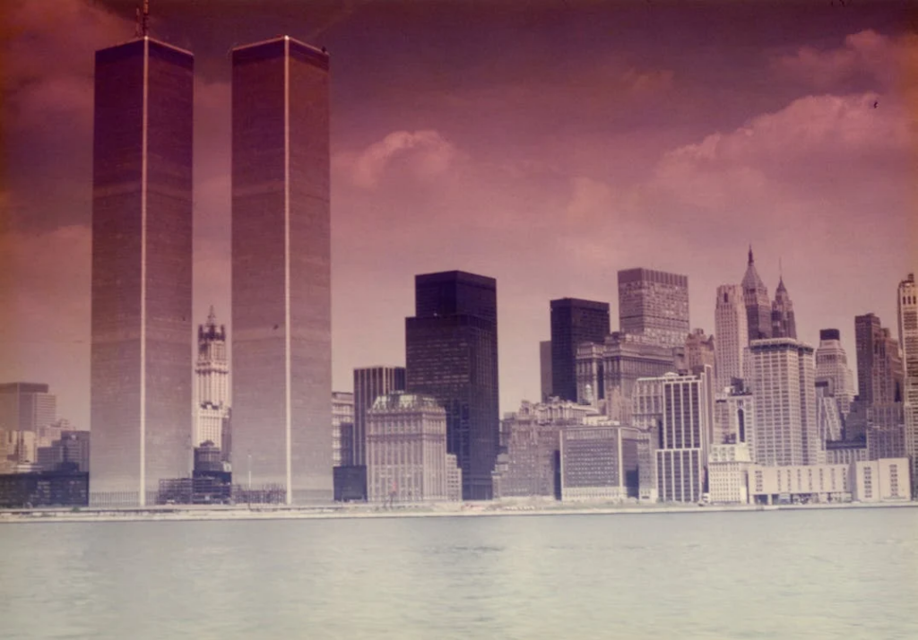 A historical photograph of the New York City skyline featuring the Twin Towers of the World Trade Center before the 9/11 attacks. The iconic twin skyscrapers dominate the left side of the image, standing tall against a purple-tinted sky. The Manhattan skyline stretches across the harbor, with water visible in the foreground.