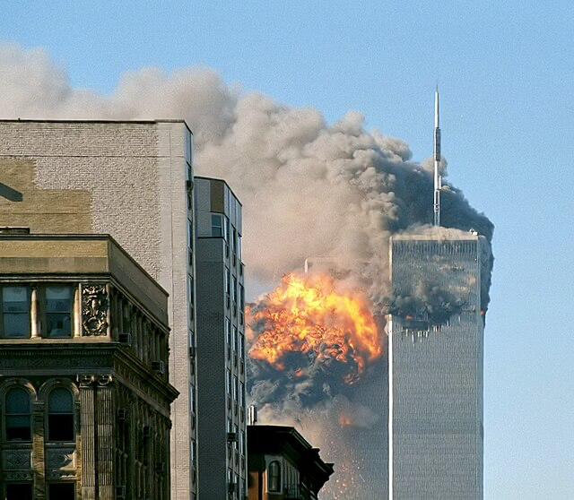 A still from rare 9/11 footage capturing the exact moment of impact when the second plane hit the South Tower of the World Trade Center on September 11, 2001. The image shows a large fireball and black smoke erupting from the tower against a clear blue sky. The North Tower, already damaged and smoking, is visible beside it. The photo is taken from nearby buildings in Manhattan, showing the horrific moment from a street-level perspective that few cameras captured with such clarity.