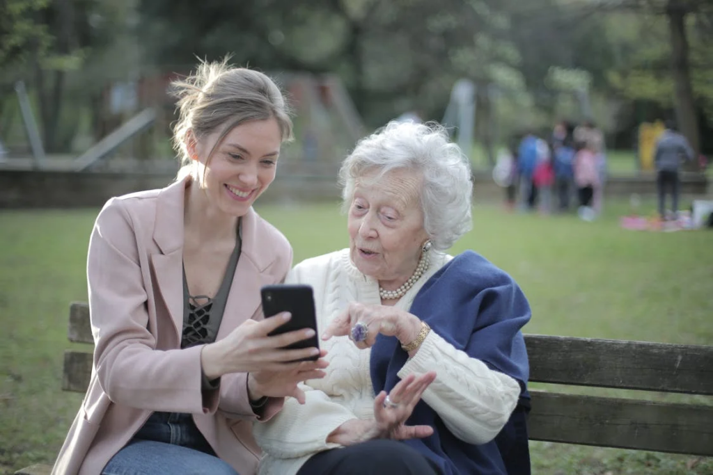 A young woman in a light pink blazer sitting on a park bench with an elderly woman wearing a blue cardigan and pearl necklace. The younger woman is showing something on her smartphone to the older woman, who appears engaged and interested. This image represents intergenerational connection and support, which is vital throughout the dementia journey.