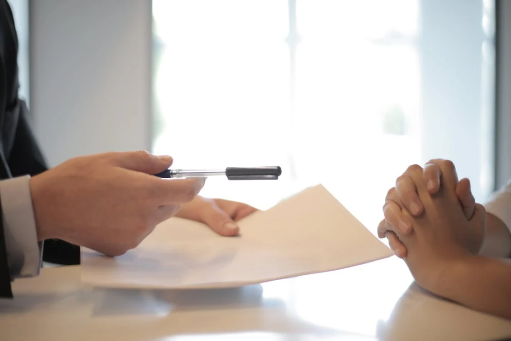 This image shows a close-up of two people's hands at a table, possibly representing a legal consultation regarding a sex offender case. On the left, someone in professional attire (visible sleeve of a suit or jacket) is holding a pen and pointing to a document on the table. On the right side are another person's clasped hands. The setting appears to be a formal meeting or legal discussion, with bright natural light coming through a window in the background.