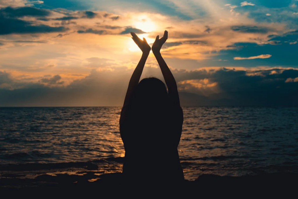 Silhouette of person with raised arms against sunset over ocean, symbolizing hope and resilience in the face of challenges.