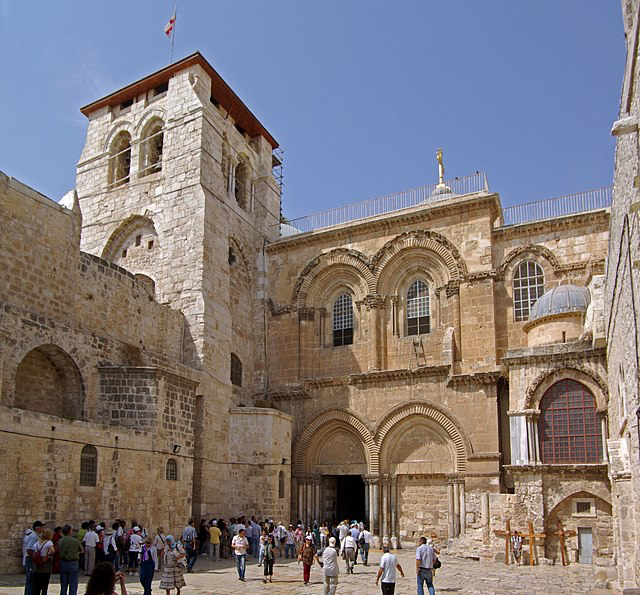 Exterior view of the Church of the Holy Sepulchre in Jerusalem, the historic structure housing Jesus' burial site. The ancient stone building features Roman and Byzantine architectural elements with arched entrances. Visitors can be seen walking in the courtyard approaching the main entrance of this sacred location where archaeological discoveries have revealed new insights about Jesus' death and resurrection.