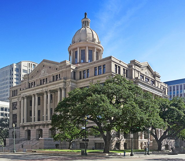 This image shows a grand government building or courthouse with classical architecture. It features a prominent dome on top, stone columns at the entrance, and ornate design elements typical of civic buildings. The structure is photographed against a blue sky, with mature trees in the foreground. It appears to be a county courthouse or similar government facility, representing the judicial system.