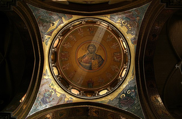 Interior dome of the Church of the Holy Sepulchre showing a stunning Byzantine mosaic. The central roundel features an image of Christ Pantocrator (Christ Almighty) surrounded by intricate decorations and religious imagery. Beams of natural light illuminate the sacred space, creating a spiritual atmosphere within this ancient church.