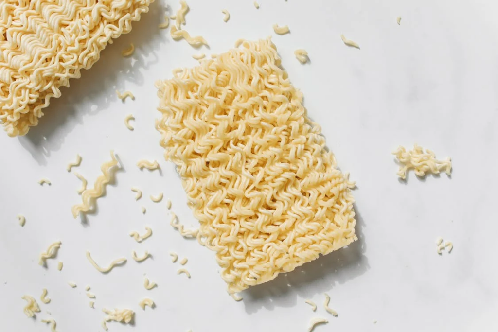 Dry instant ramen noodles broken into pieces on a white background, showing the characteristic wavy texture of processed ramen noodles.