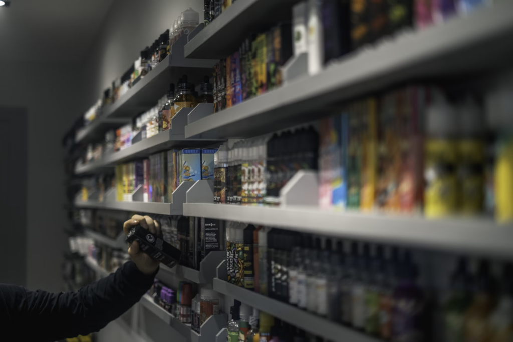A hand reaches for a colorful disposable vape on store shelves stocked with numerous vaping products, highlighting the easy accessibility that concerns many parents.