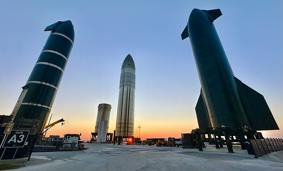 Three Starship rockets stand upright at the Starbase site. The sun is setting in the background. 