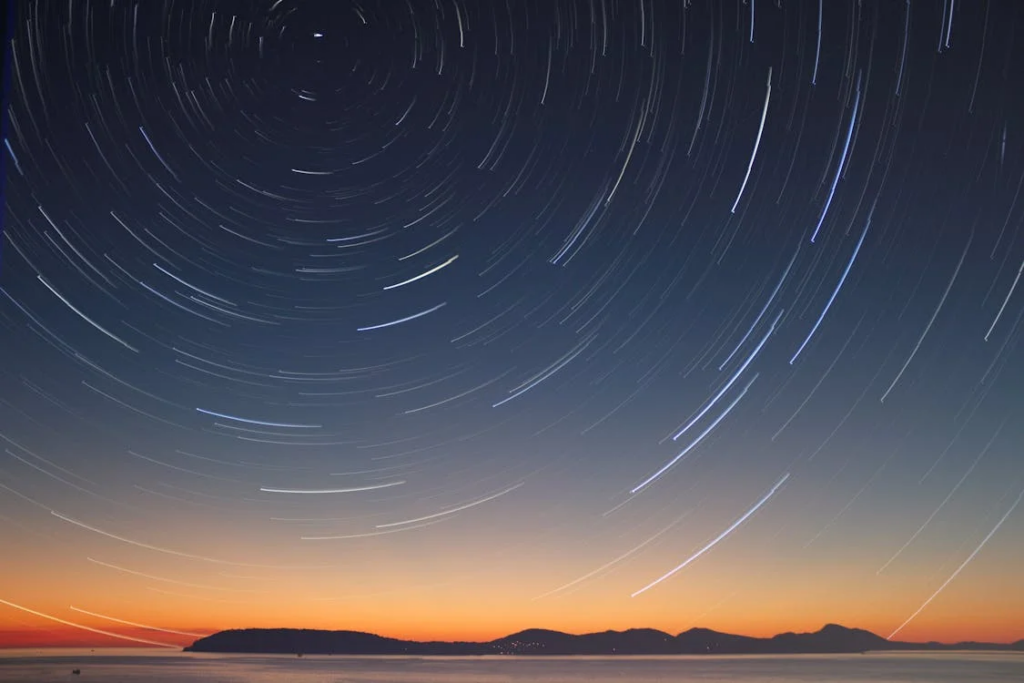 A time-lapse photograph of star trails in the night sky that would leave any flat earther speechless. The image shows concentric circular light trails against a dark blue sky gradient. The stars appear to rotate around a central point (likely Polaris, the North Star). At the bottom of the image is a silhouette of mountains or islands on the horizon with an orange-red sunset/sunrise glow. The circular pattern of the star trails demonstrates Earth's rotation.