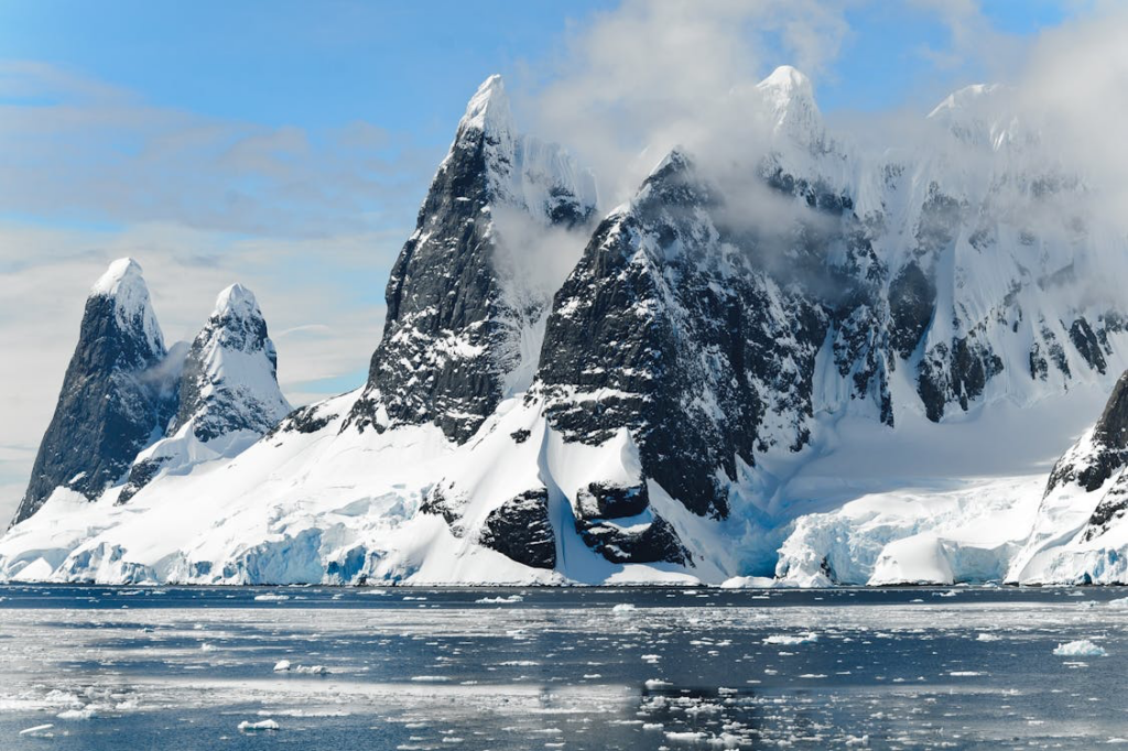A dramatic photograph of Antarctica's landscape showing steep, jagged mountains covered in snow and ice. Several tall, triangular mountain peaks rise sharply against a partly cloudy blue sky. At the base of the mountains is an ice shelf that meets the ocean, where small pieces of sea ice can be seen floating on the deep blue water. The scene captures the harsh, majestic beauty of the Antarctic wilderness.