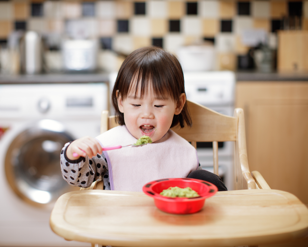Baby girl eating mashed avocado at home kitchen