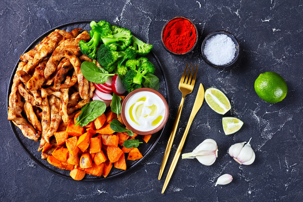 Weight loss plan: grilled chicken breast and baked sweet potato, boiled broccoli on a black plate on a dark concrete background with sour cream, radish, and spinach, garlic and lime, flat lay
