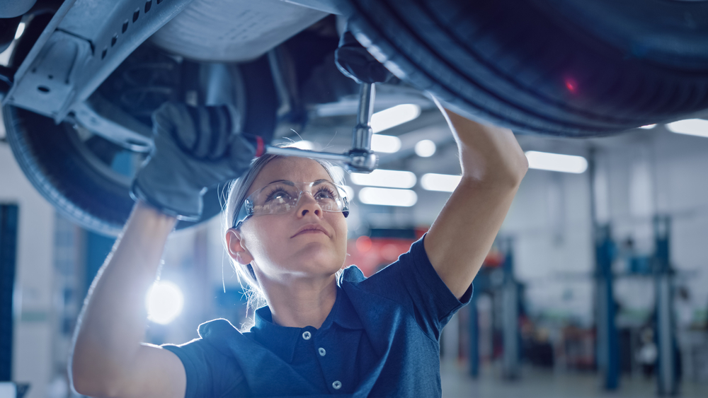Portrait Shot of a Female Mechanic Working Under Vehicle in a Car Service. Empowering Woman Wearing Gloves and Using a Ratchet Underneath the Car. Modern Clean Workshop.
