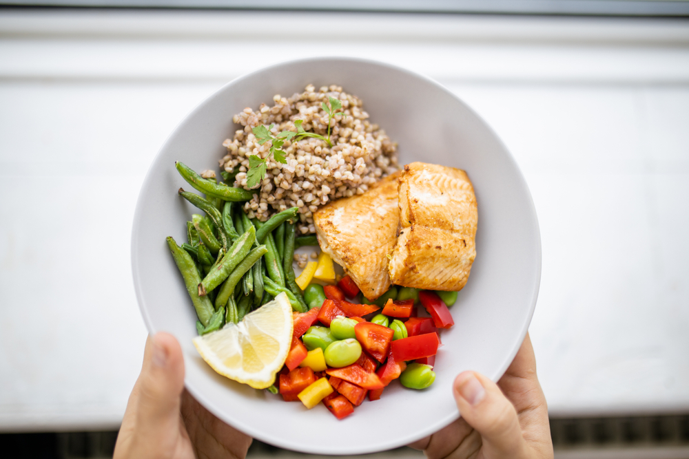 Hands holding salmon and buckwheat dish with green beans, broad beans, and tomato slices. Nutritious dish with vegetables and fish from above. Healthy balanced diet