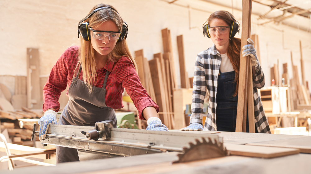 Two young women as carpenter apprentices work on the circular saw in the workshop

