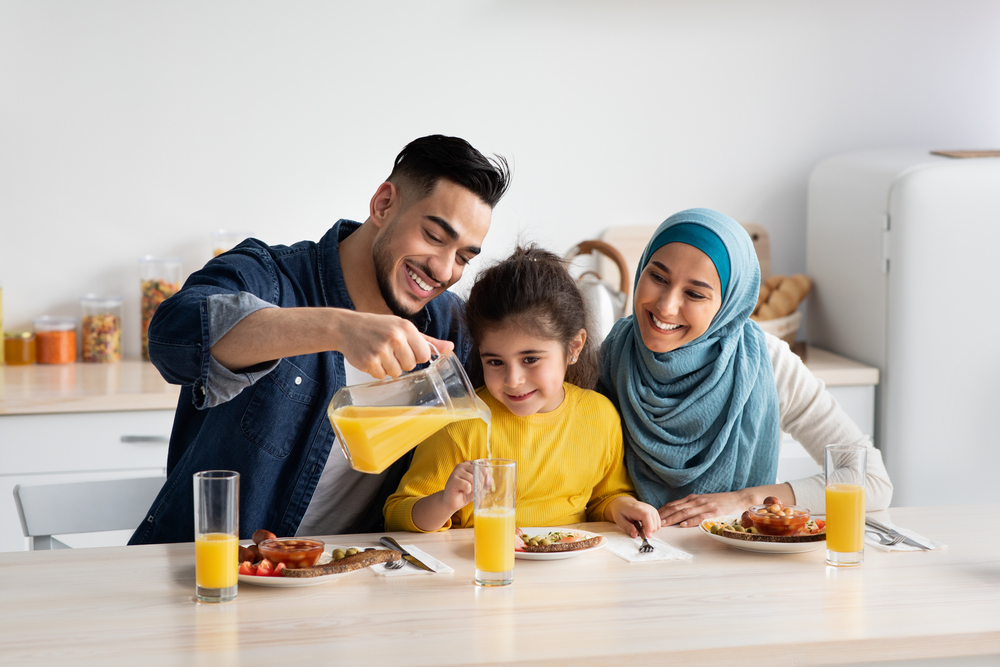 Portrait Of Happy Middle Eastern Family Of Three Having Breakfast In Kitchen Together, Arab Parents With Cute Little Daughter Enjoying Tasty Food And Drinking Orange Juice While Sitting At Table

