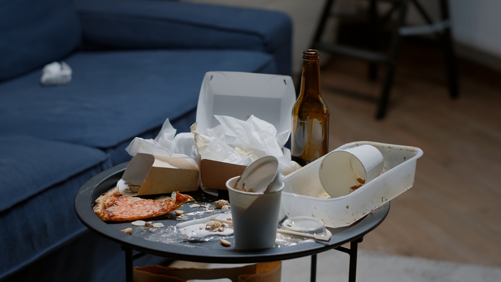 Close up of messy table, leftover food, spilled drinks, dirty dishes in empty living room, bottle of beer and napkins on blue sofa. Unorganized apartment of person with sever depresion having rubbish