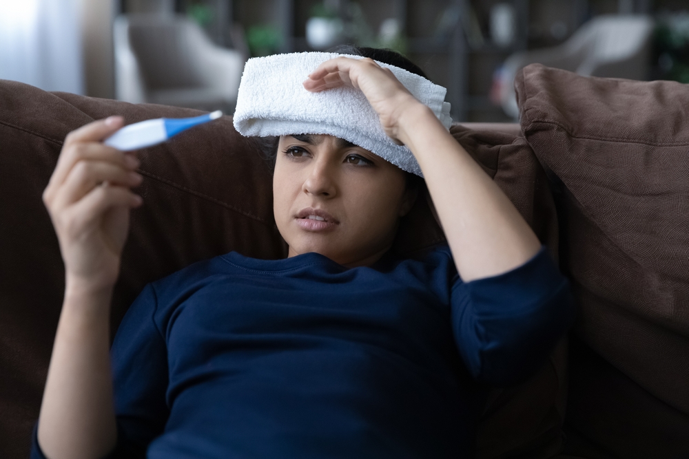 Unhealthy young Indian woman measuring body temperature with electronic thermometer, holding wet towel compress on forehead, suffering from severe headache or first flu grippe covid19 symptoms.