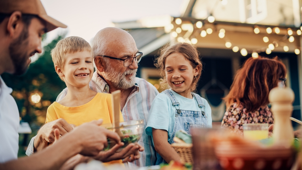 Happy Senior Grandfather Talking and Having Fun with His Grandchildren, Holding Them on Lap at a Outdoors Dinner with Food and Drinks. Adults at a Garden Party Together with Kids.
