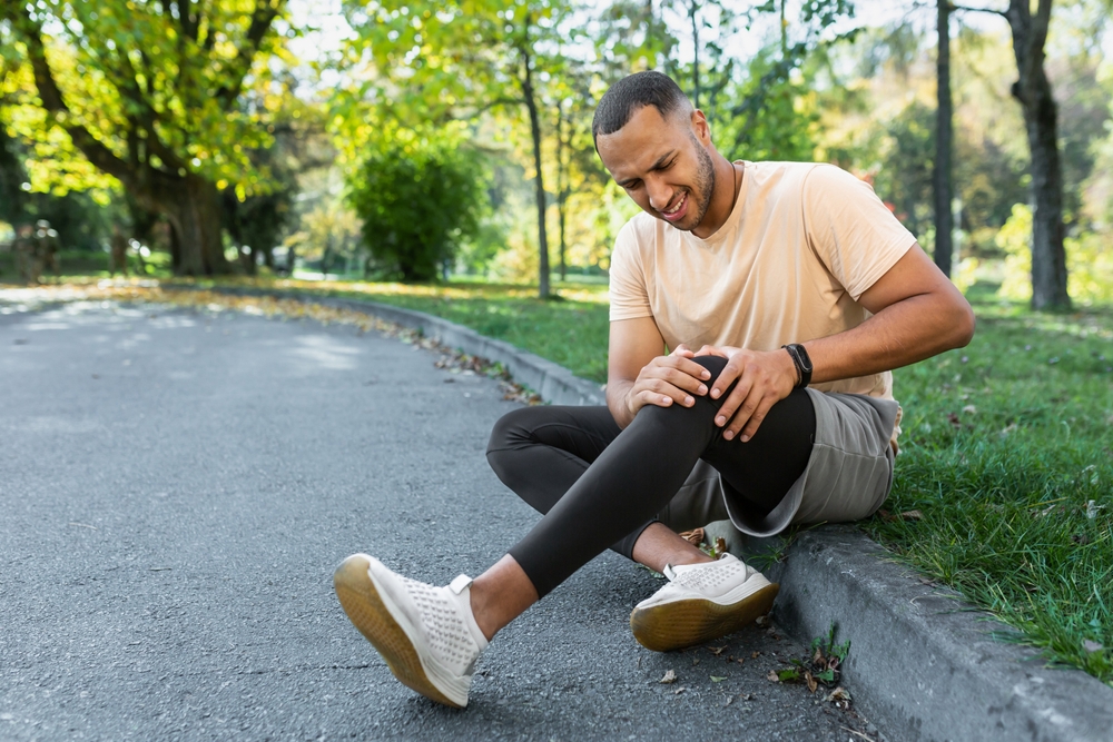 Man injured leg while jogging, african american man sitting on ground, massaging sore muscle with hands, upset and injured runner.