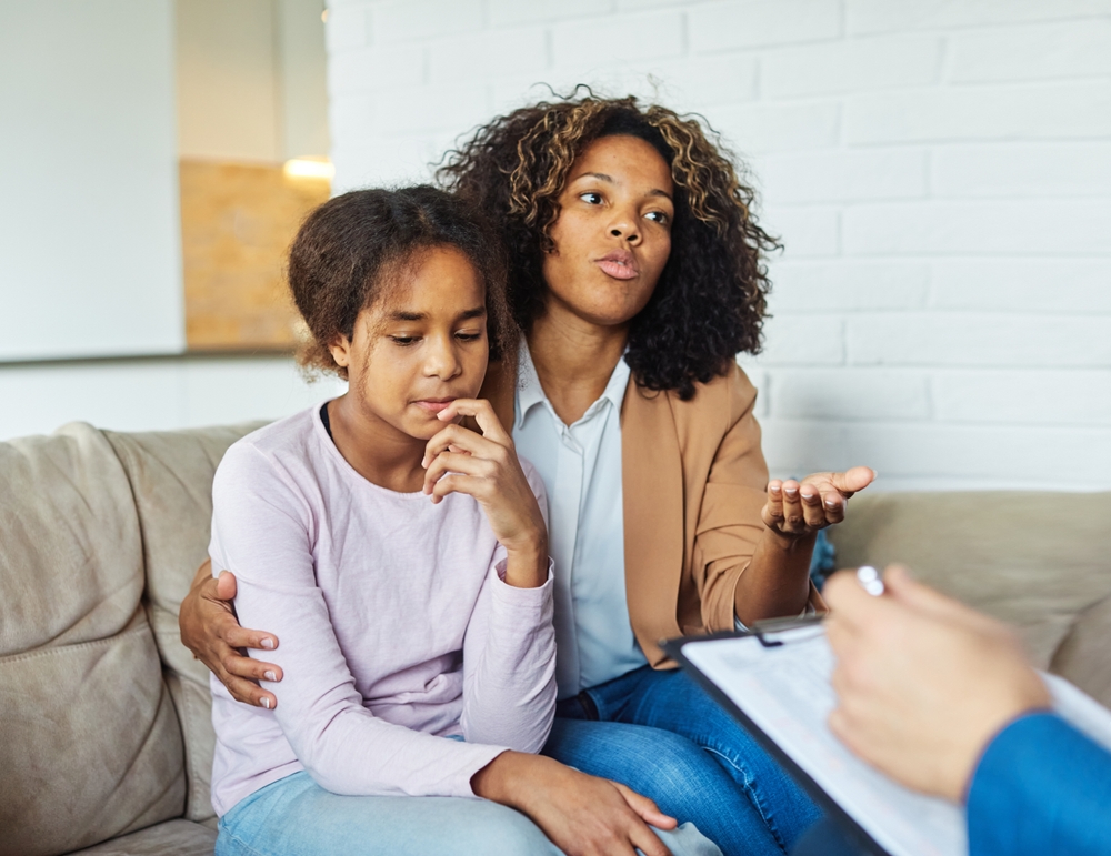 Mother with her teenage daughter at meeting with social worker, psychologist discussing mental health family sitting on sofa in psychotherapist office
