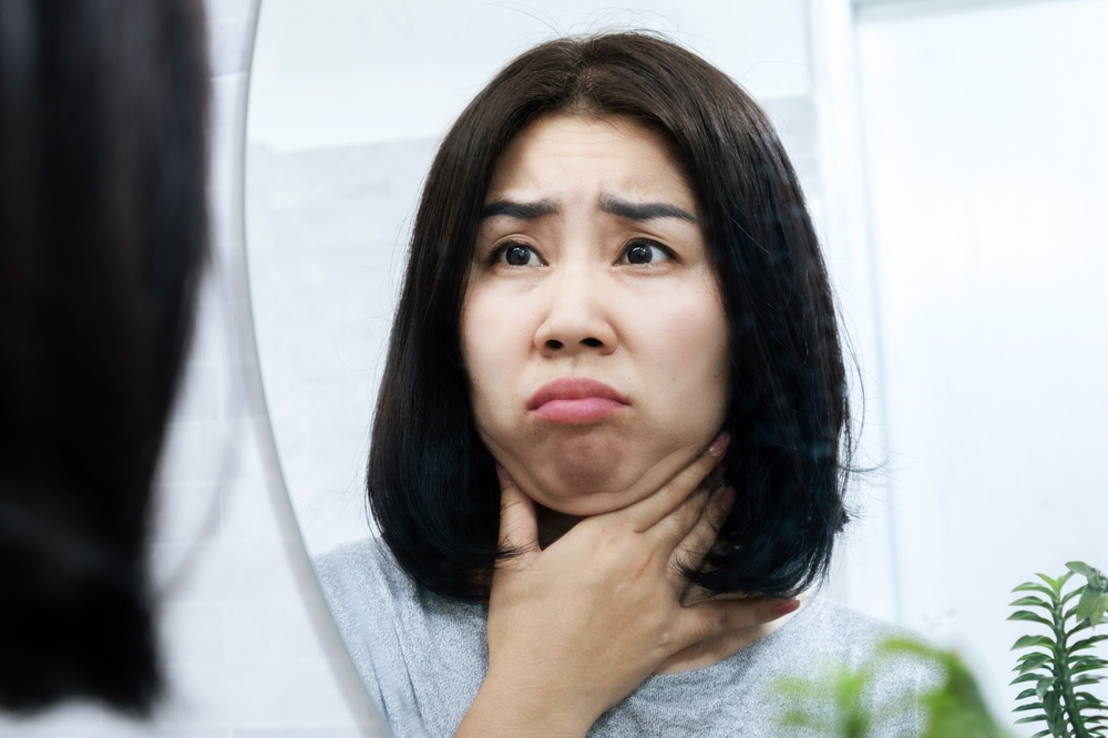 worried Asian woman checking her double chin under her lower jaw in front of a mirror, a sign of weight gain or obesity
