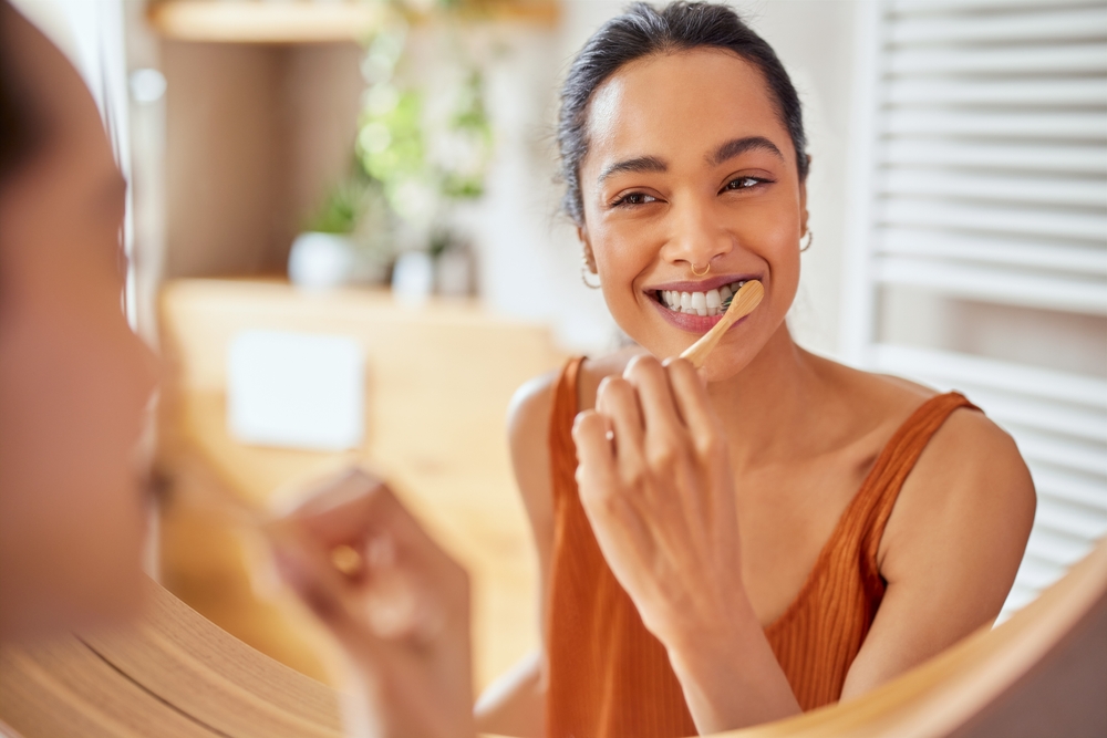 Smiling young woman brushing teeth in bathroom. Girl looking in mirror while using ecological toothbrush with whitening toothpaste to clean her teeth. Multiethnic girl cleaning teeth in the morning.
