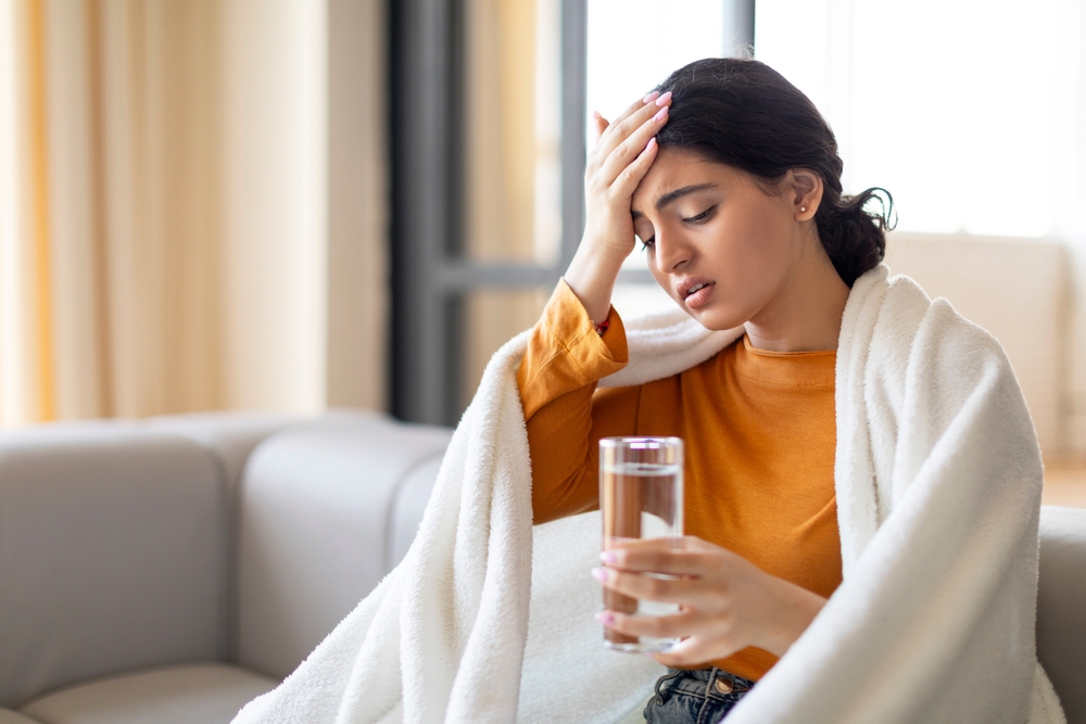 Headache Concept. Sick Young Indian Female Suffering From Migraine At Home, Ill Eastern Woman Holding Glass Of Water, Touching Forehead And Frowning, Feeling Fever, Closeup Shot With Copy Space