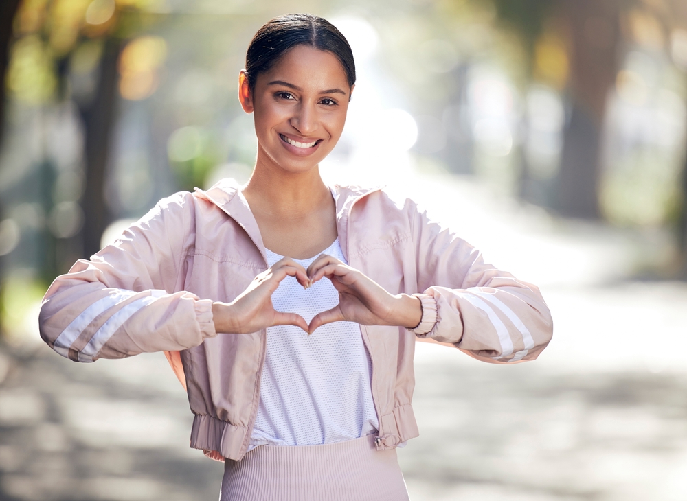 Girl, portrait and outdoor for fitness, heart and sportswear in nature, smile and exercise on road. Wellness, city and runner with happiness, hands and gesture of love, healthy and bokeh in park
