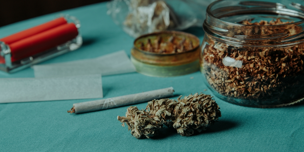 closeup of a cannabis bud on a table next to a used herb grinder and a jar with some rolling tobacco, in a panoramic format to use as web banner or header
