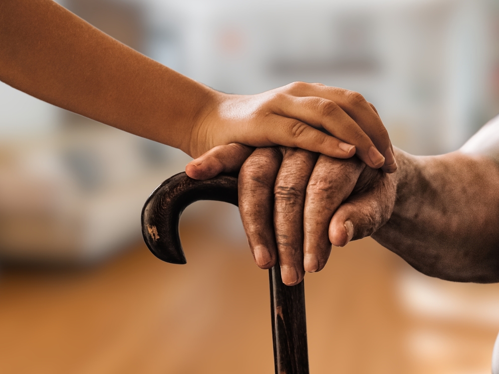An elderly person holding a cane with a younger hand offering support, symbolizing assistance and stability for senior citizens.
