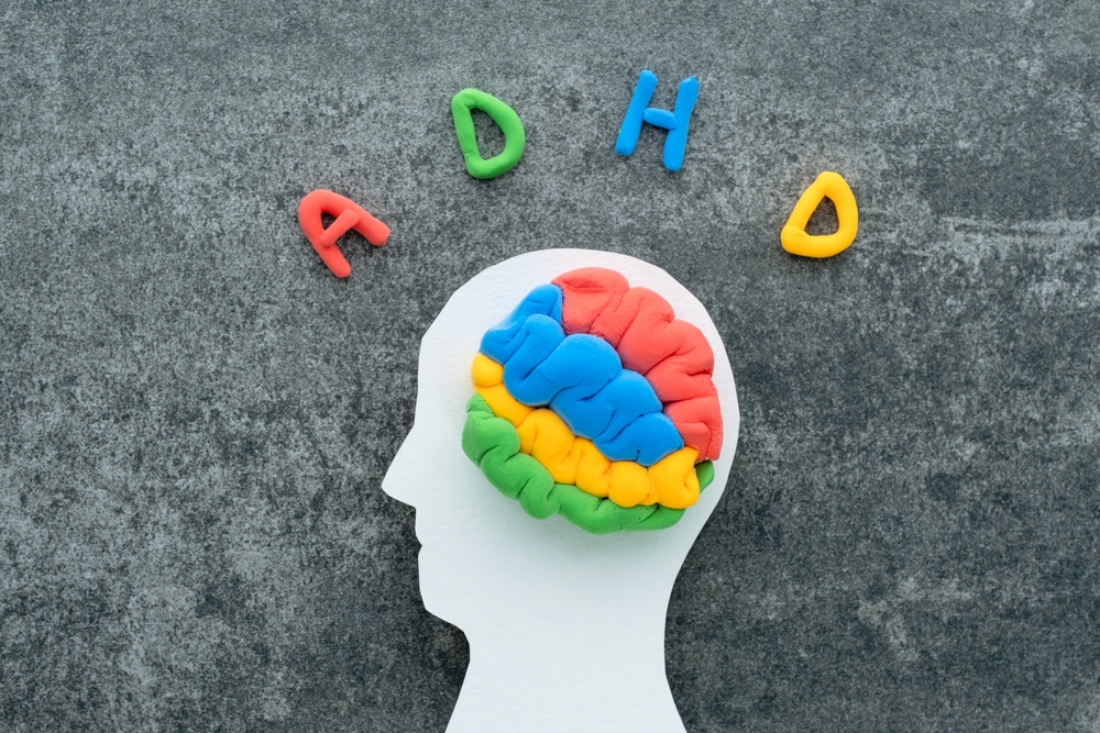 A brain model crafted from colorful materials rests on a white head silhouette, with the letters ADHD on a concrete background. ADHD and learning difficulties awareness and support
