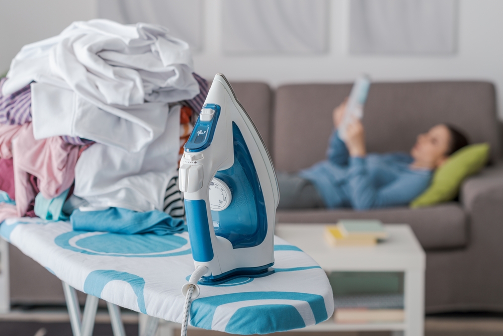Lazy careless housewife on the couch at home and a pile of laundry on the ironing board on the foreground, boring household chores concept