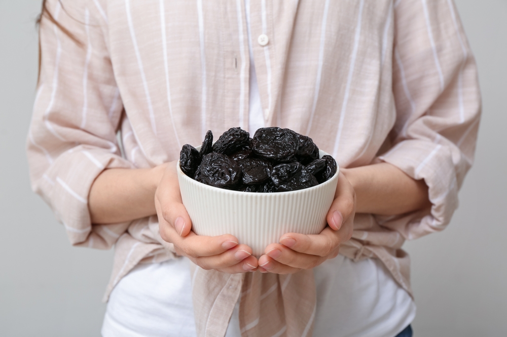 Woman holding bowl with dried prunes on grey background, closeup
